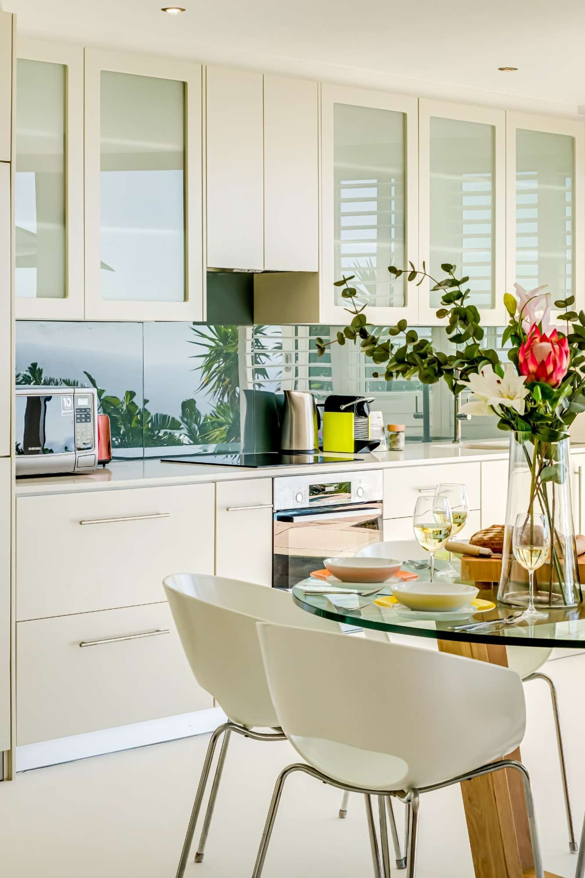 A modern kitchen with a glass table, white chairs, and a view of the outdoors.