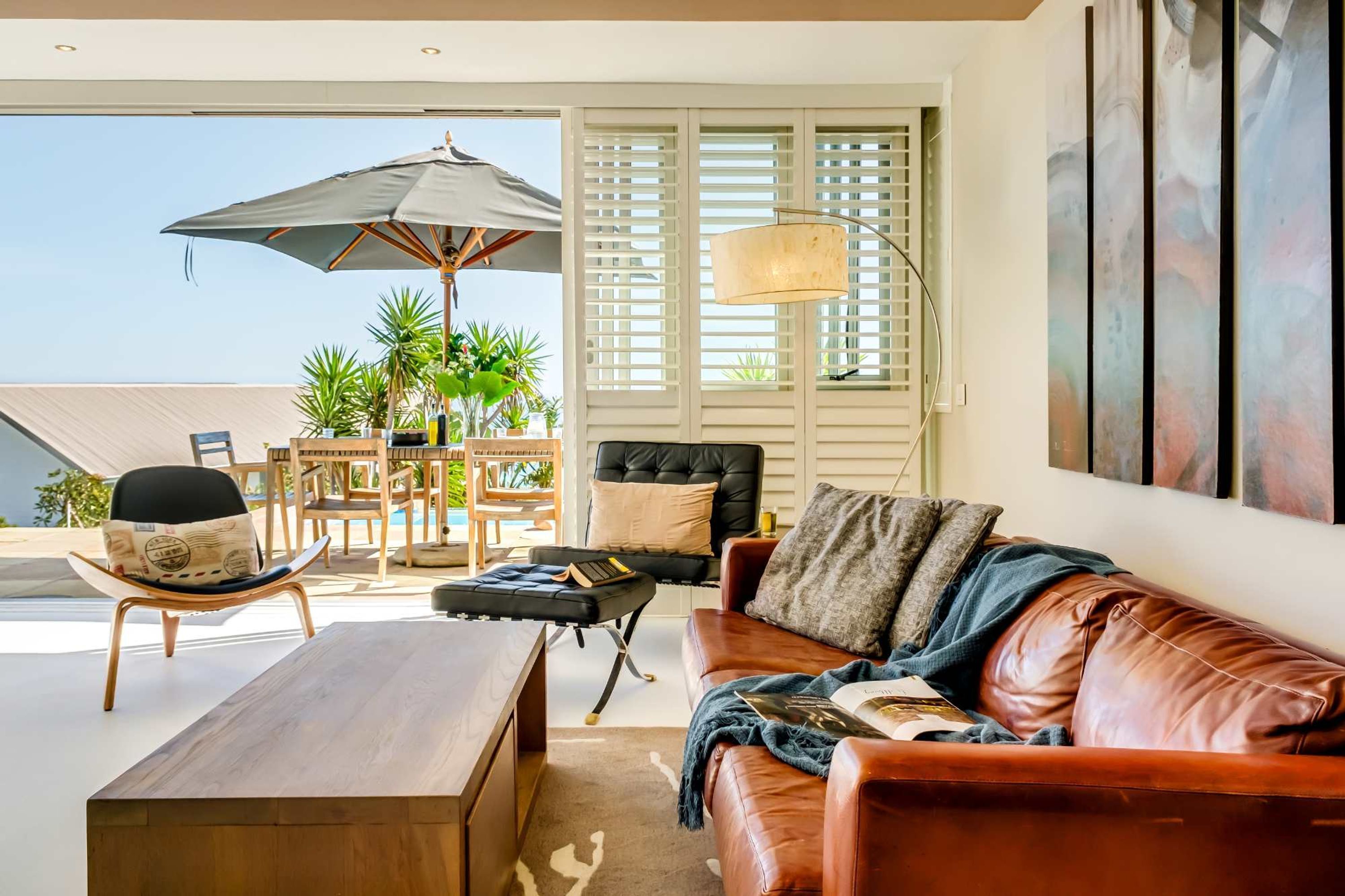 A living room with a brown leather sofa, a wooden coffee table, a black chair, a wooden dining table with chairs, a large umbrella, and a window with blinds.