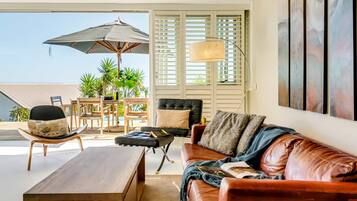 A living room with a brown leather sofa, a wooden coffee table, a black chair, a wooden dining table with chairs, a large umbrella, and a window with blinds.