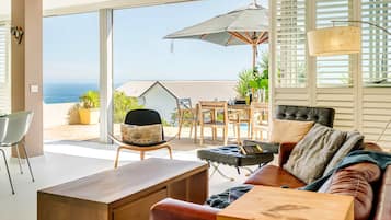 A modern living room with a view of the ocean, a wooden desk, a leather sofa, and a dining area with outdoor furniture.