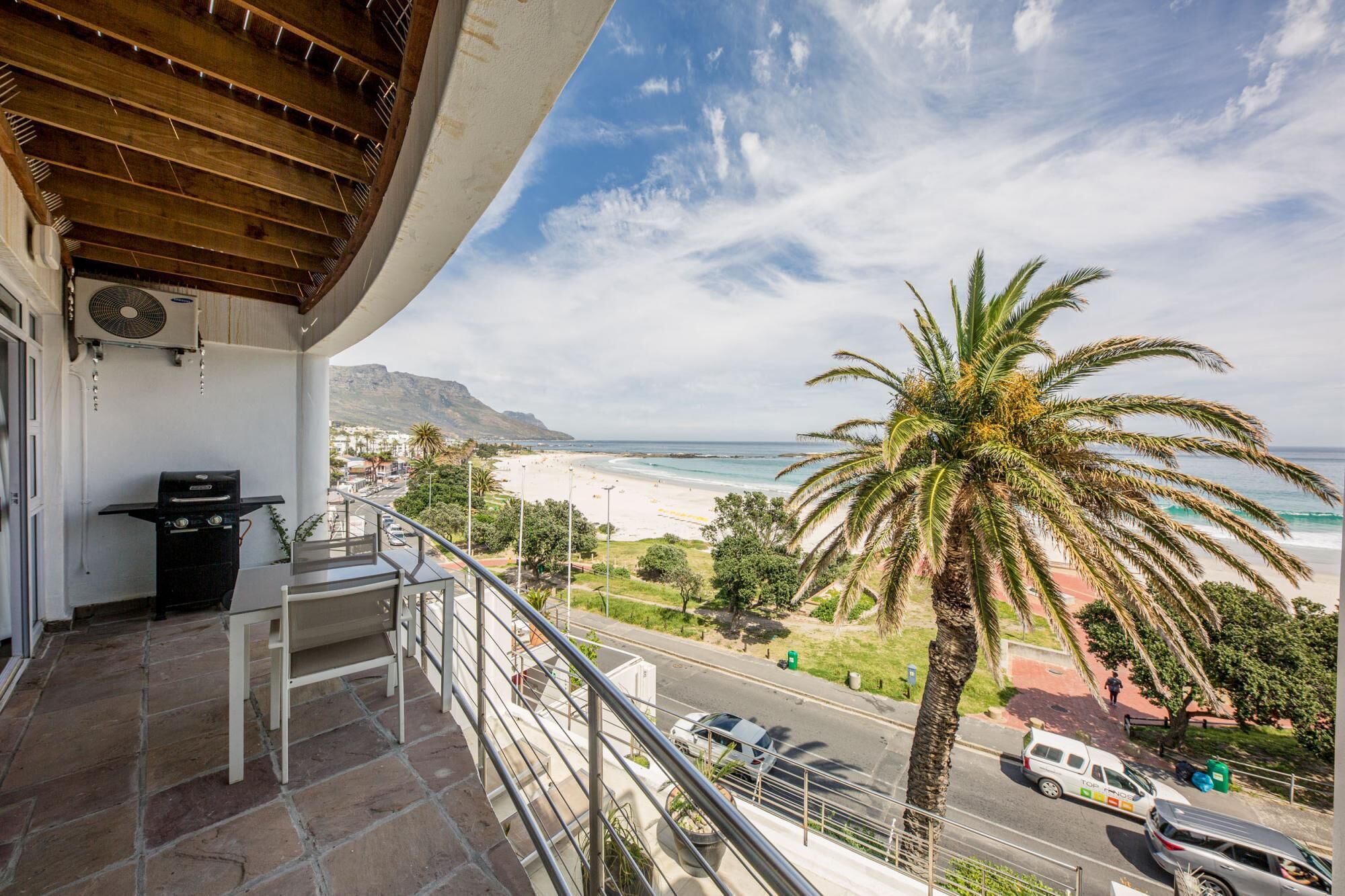 A balcony with a grill, a view of the beach, and a palm tree.