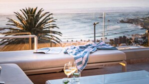 A sunlit balcony with a view of the ocean, a lounge chair, a glass table with drinks, and a striped towel. - Spectacular Holiday Villa w Private Pool Gym Prima Views (Cape Town)