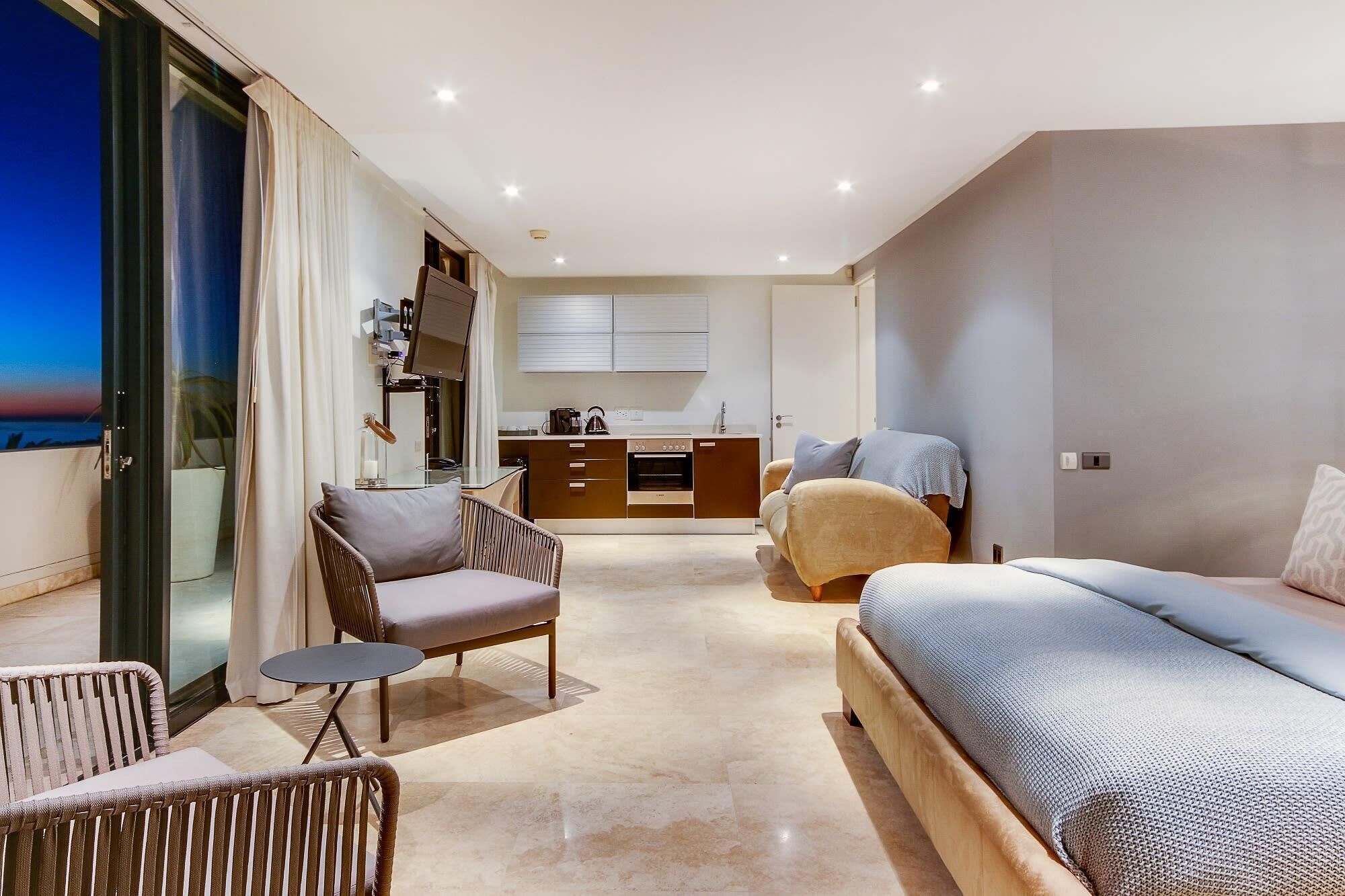 A modern bedroom with a bed, a sofa, a chair, and a small table. There is a kitchenette in the background with cabinets and a countertop.