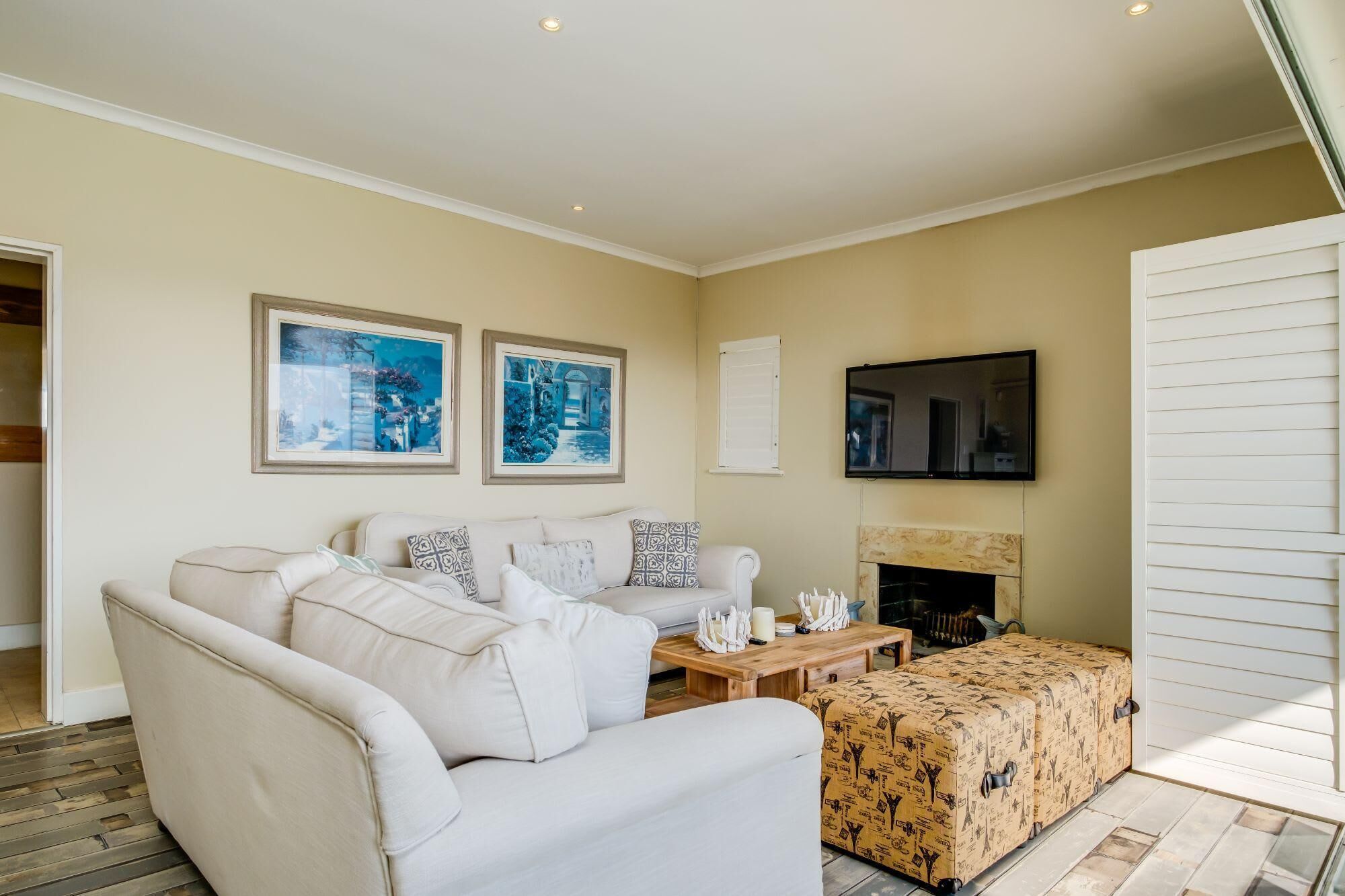 Een woonkamer met een beige bank, een ottoman met patroon, een houten salontafel en een open haard. Twee ingelijste foto's hangen aan de muur en er is een flatscreen televisie boven de open haard bevestigd.