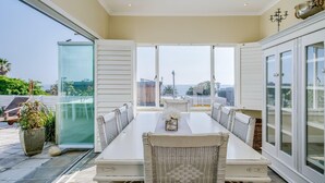 A dining area with a long table and wicker chairs, large windows offering a view of the sea and buildings, and a glass door leading outside.