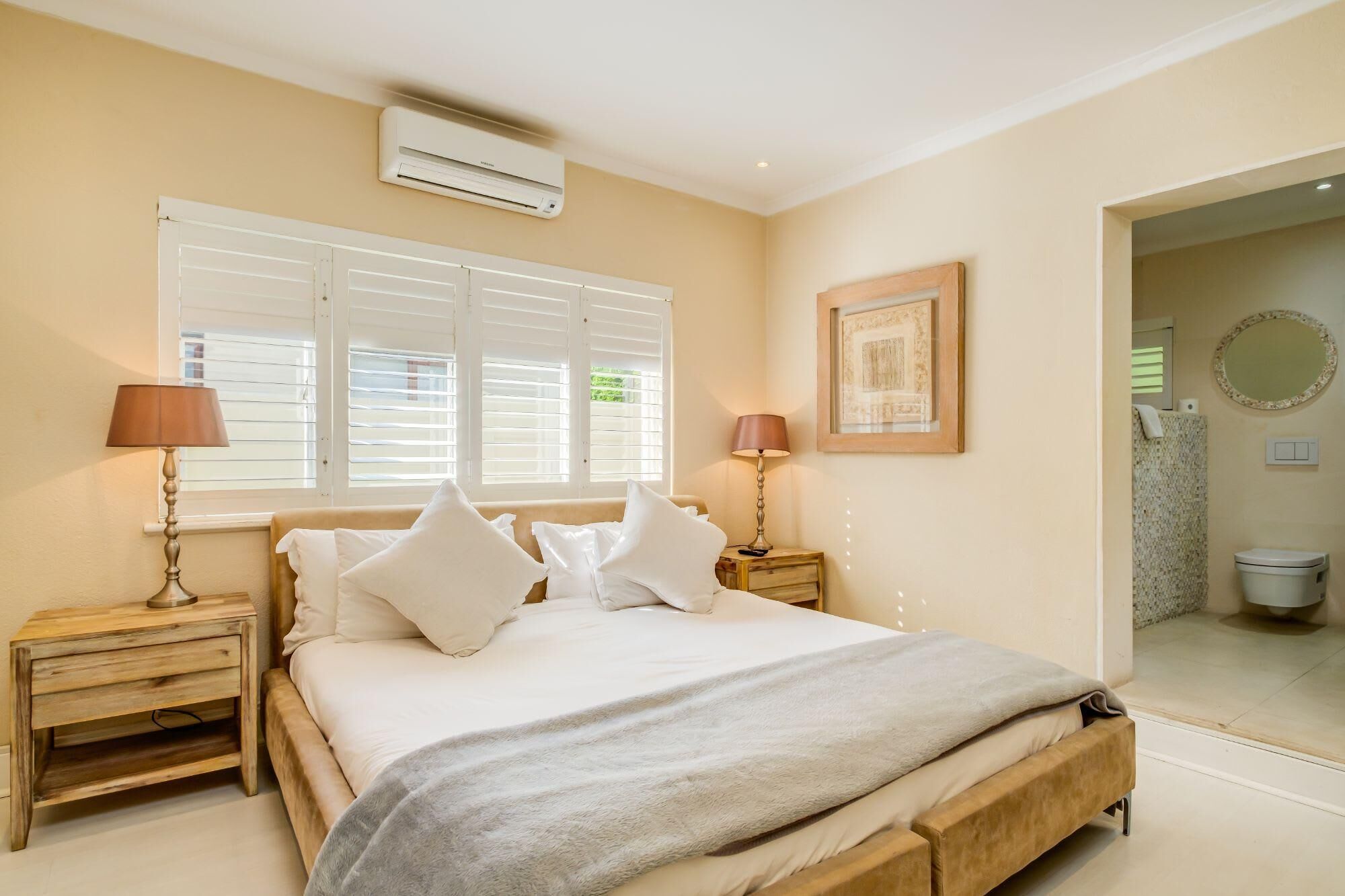 A bedroom with a bed, two bedside tables, a lamp, a window with blinds, an air conditioning unit, a framed picture, and a bathroom visible through an open door.