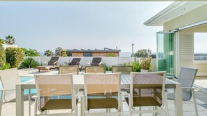 A sunlit outdoor seating area with white chairs and a pool, overlooking a modern building with a view of the sea.