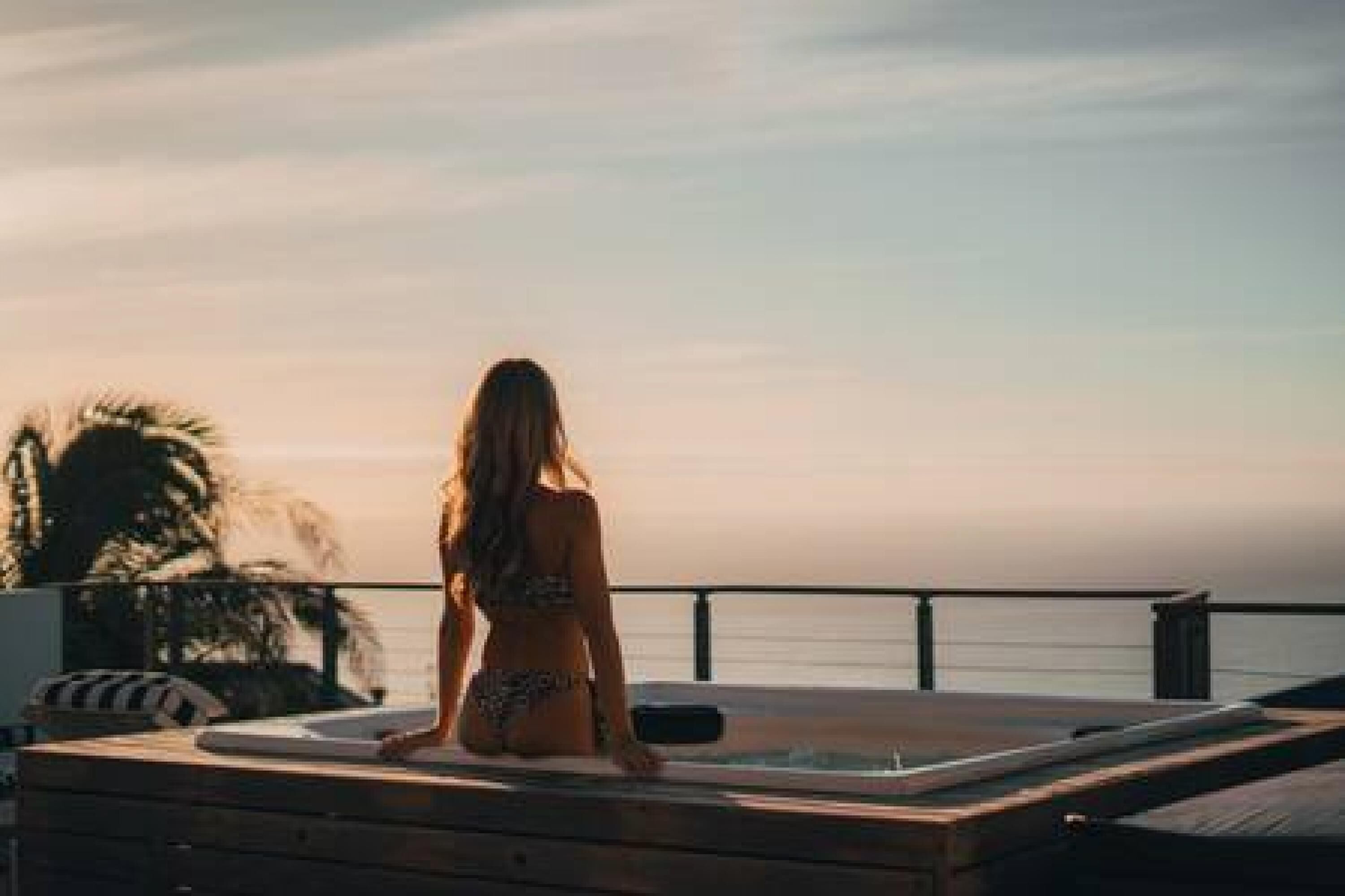 Une femme est assise sur une terrasse en bois donnant sur une étendue d’eau, avec un jacuzzi et des chaises longues visibles.