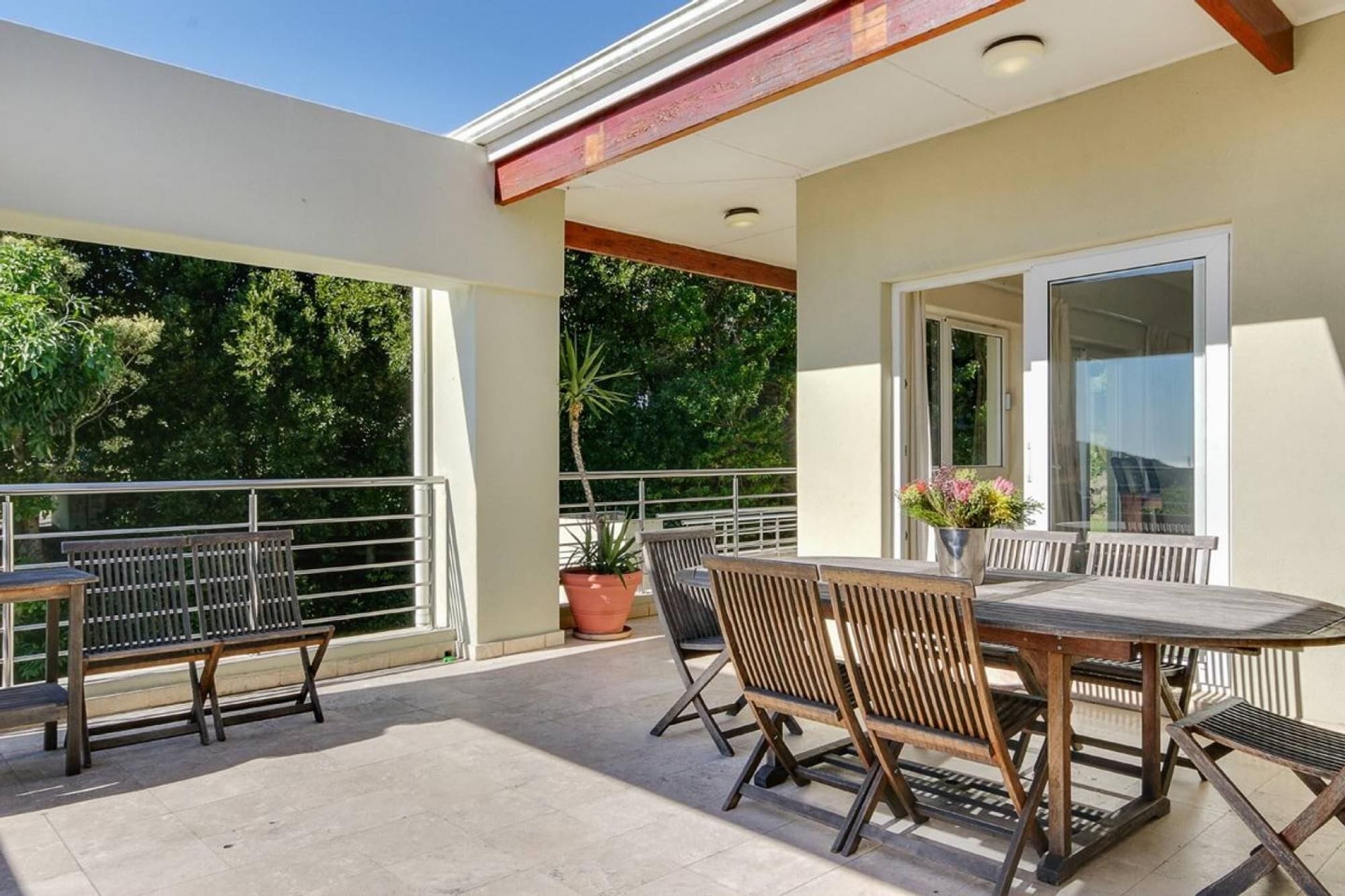 A covered outdoor patio with a table and chairs, potted plants, and a view of trees.