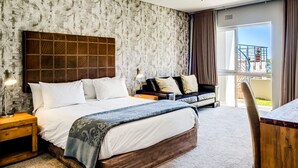 A hotel room with a large bed, a wooden headboard, a patterned wall, a balcony with a view, and a dining area with a table and chairs. - Family Home With Views Pool Garden 100 Geneva (Cape Town)
