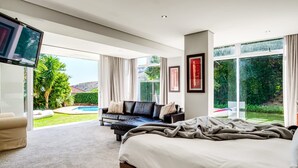 A spacious bedroom with a large bed, a sofa, and a TV. It features large windows and a view of a pool and garden. - Family Home With Views Pool Garden 100 Geneva (Cape Town)