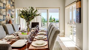 A dining room with a long table set for a meal, a vase with flowers, and a view of the ocean through large windows. - Family Home With Views Pool Garden 100 Geneva (Cape Town)