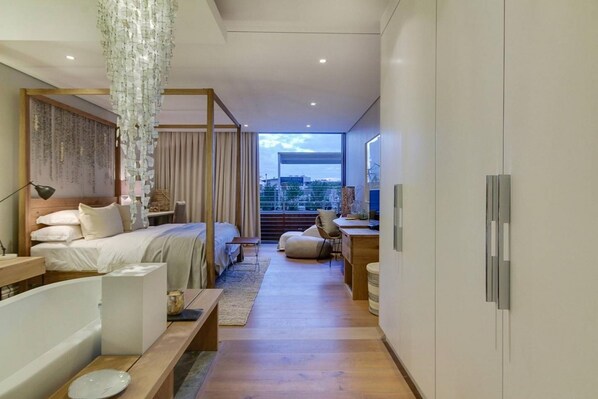 A modern bedroom with a large bed, bedside tables, a desk, and a view of the city.