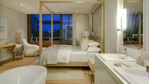 Une chambre d’hôtel moderne dotée d’un grand lit, d’un coin salon séparé avec un fauteuil, d’une grande fenêtre et d’une salle de bain avec une baignoire et un lavabo.