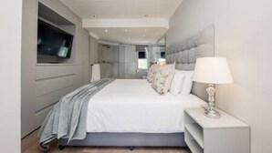A modern bedroom with a large bed, a TV mounted on the wall, and a bedside table with a lamp. - Holiday Apartment Opposite the Beach Seasonsfind the Sunset (Cape Town)