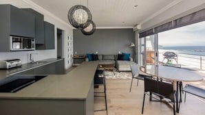 A modern kitchen with a central island, a dining area with a round table, and a view of the sea through large windows. - Holiday Apartment Opposite the Beach Seasonsfind the Sunset (Cape Town)