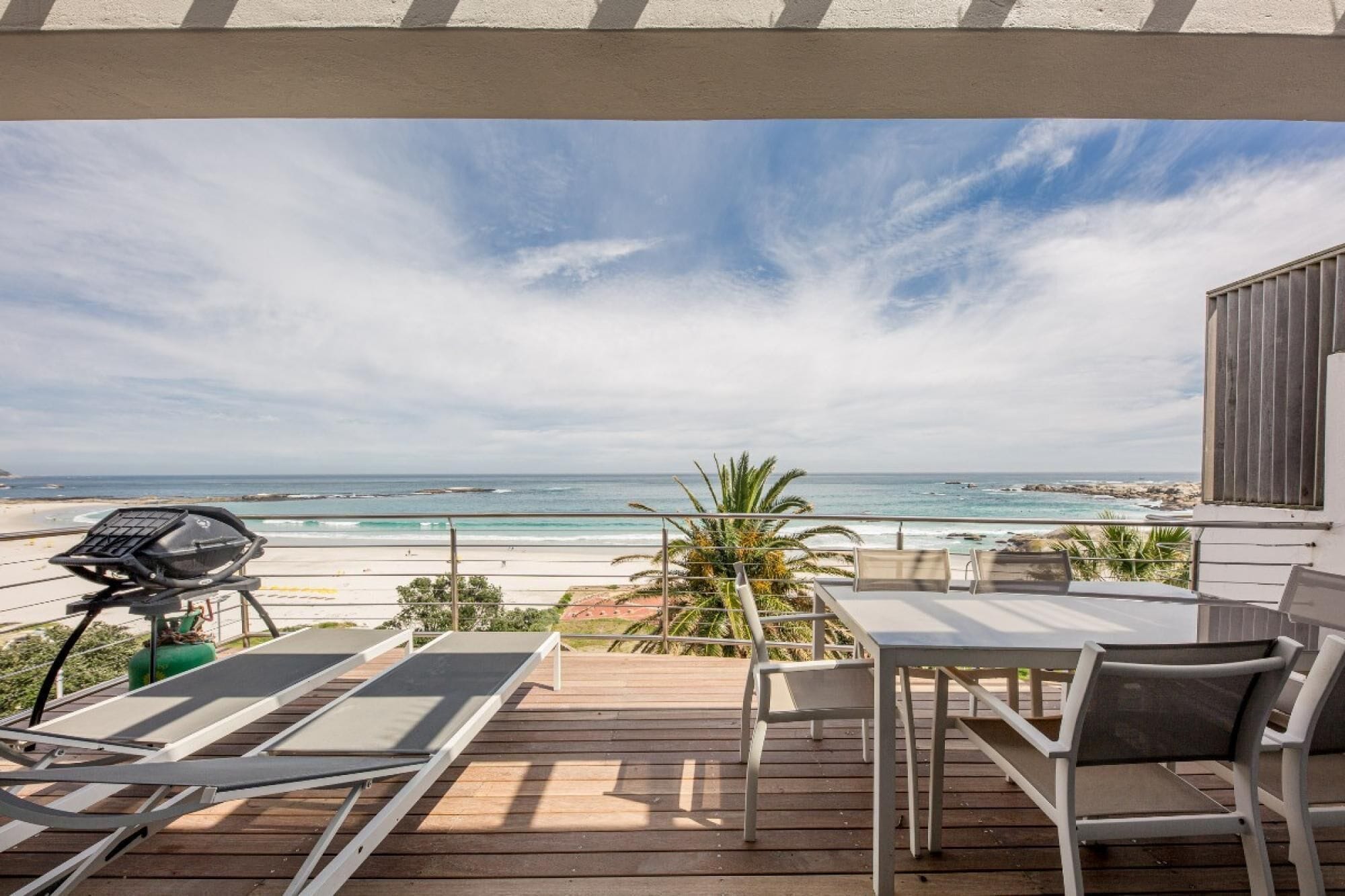 A wooden deck with a table and chairs, overlooking a beach with the ocean and rocky coastline.