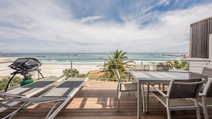 A wooden deck with a table and chairs, overlooking a beach with the ocean and rocky coastline. - Holiday Apartment Opposite the Beach Seasonsfind the Sunset (Cape Town)