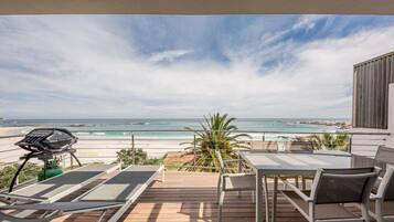A wooden deck with a table and chairs, overlooking a beach with the ocean and rocky coastline.