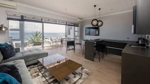 A modern living room with a sofa, coffee table, and a TV. There is a kitchen area with a bar and a large window overlooking the ocean. - Holiday Apartment Opposite the Beach Seasonsfind the Sunset (Cape Town)