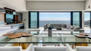 Un salon moderne avec une table basse en verre, des canapés blancs et une grande fenêtre donnant sur l’océan.