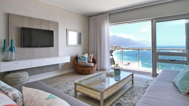 A modern living room with a flat-screen TV, a glass coffee table, a sofa, and a view of the ocean.