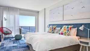 A modern hotel room with a large bed, a red chair, a round ottoman, and a view of the sea through the window.