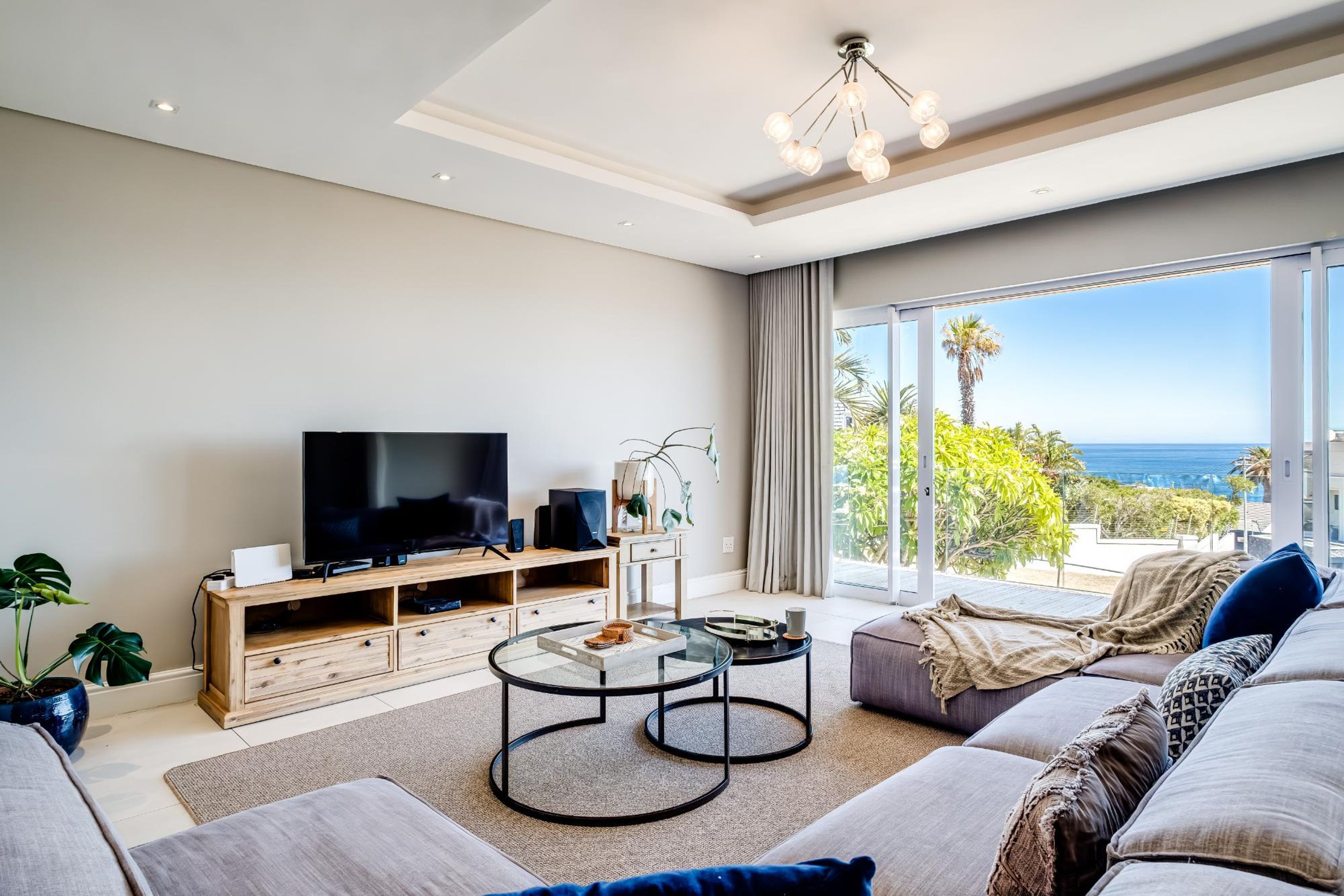 A modern living room with a large flat-screen TV, wooden entertainment unit, glass coffee tables, and a sofa with a view of the ocean.