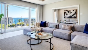 A modern living room with a large sofa, a glass coffee table, and a view of the cityscape through large windows. - Spacious Holiday Villa w Private Pool Garden Serein (Cape Town)