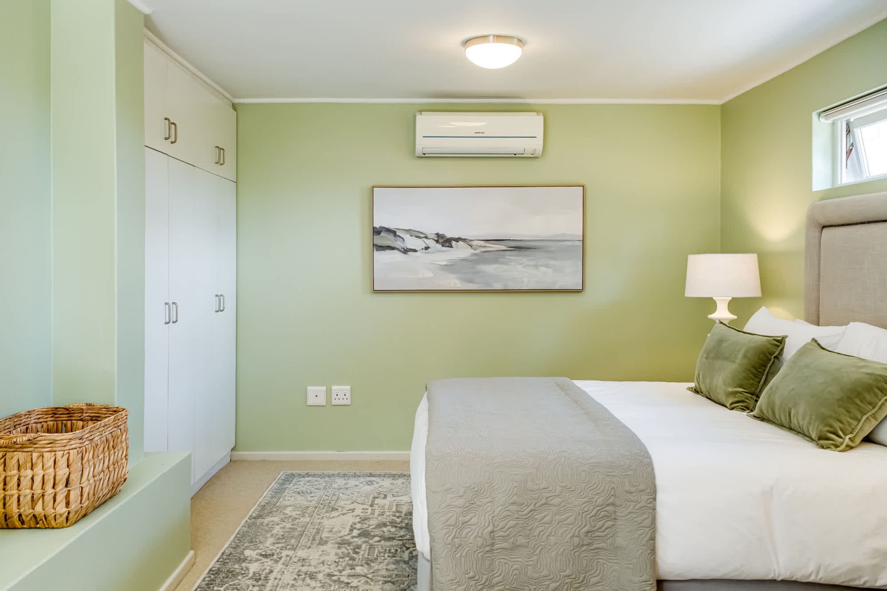 Un dormitorio con cama, lámpara, un cuadro en la pared y una cesta en el suelo.