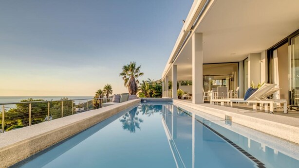 A modern pool area with a clear view of the ocean, lounge chairs, and a covered seating area. - Incredible Villa w Lap Pool Ocean Views Casa Di Sorrento (Cape Town)