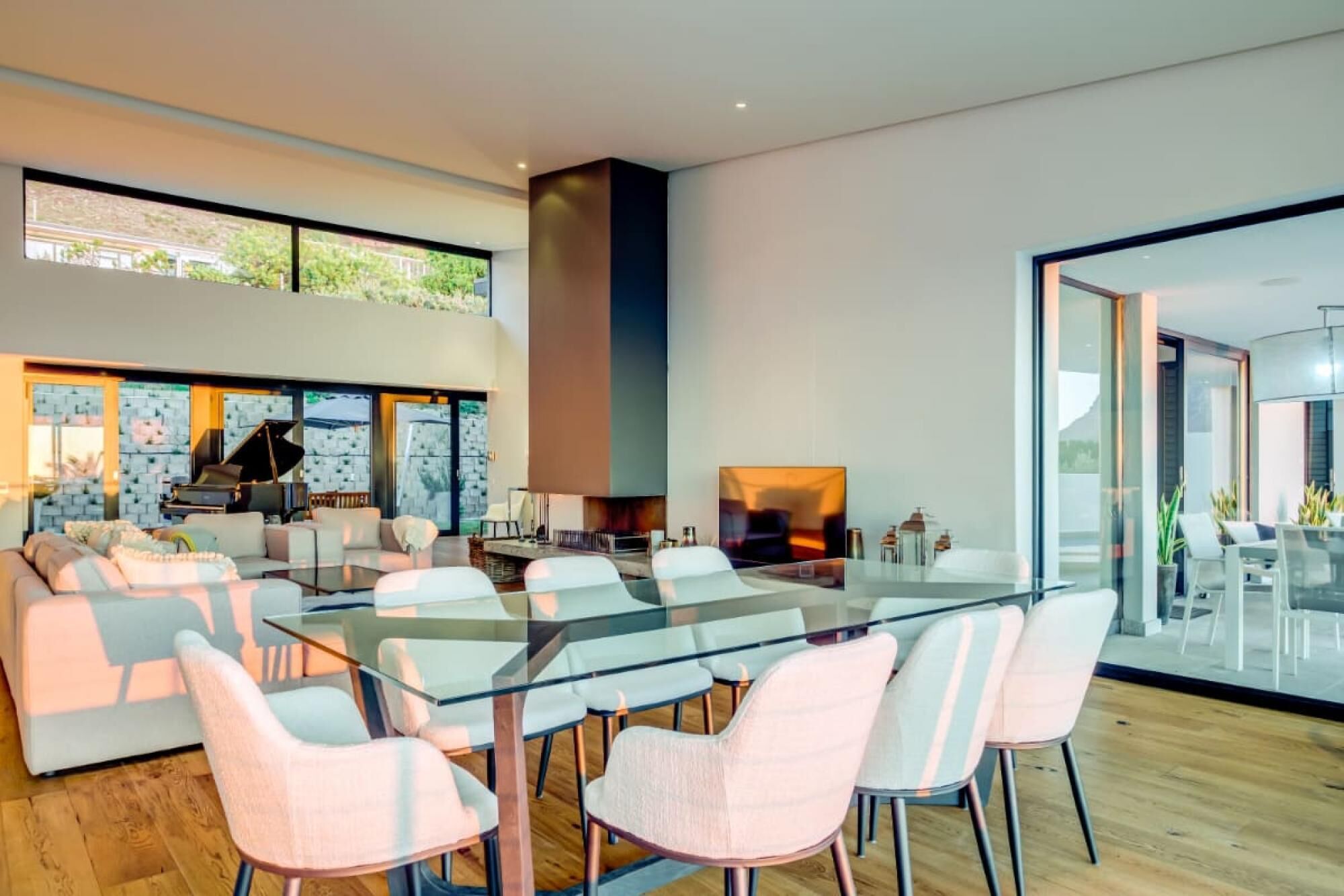 A modern living room with a glass dining table, white chairs, a piano, and a fireplace.