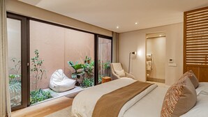 A modern bedroom with a large bed, a seating area with a chair and a small table, and a sliding glass door leading to a garden. - Stylish Apartment With Rooftop Deck Pool 53 Napier (Cape Town)