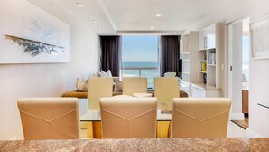 A modern dining area with a view of the ocean, a bookshelf, and a painting on the wall. - Clifton Apartment w Direct Beach Access Marella (Cape Town)