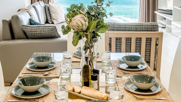 A dining area with a view of the ocean, a sofa, and a television. - Clifton Apartment w Direct Beach Access Marella (Cape Town)