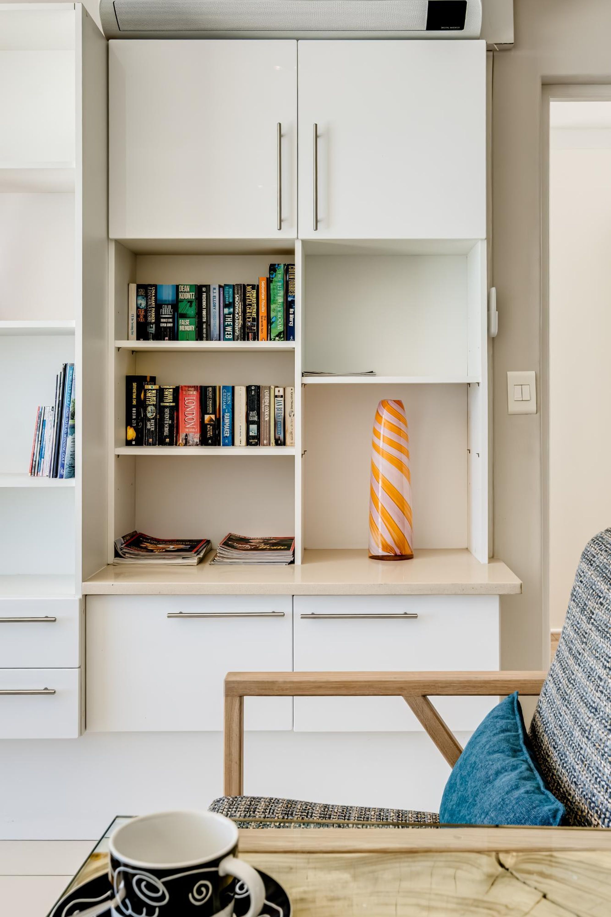 A modern living room with a white bookshelf, a wooden chair, a patterned cushion, and a coffee table with a cup and saucer.