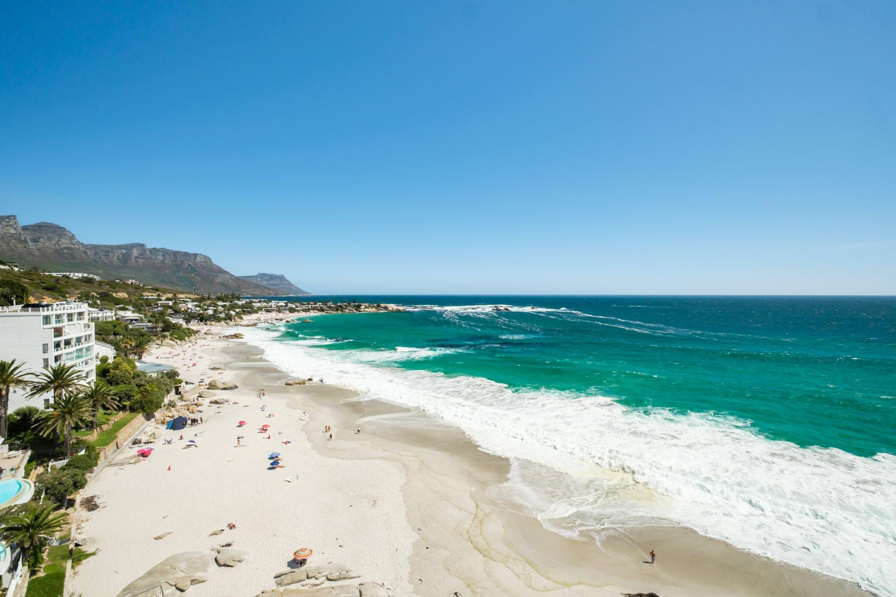 A beach with clear turquoise waters, a sandy shore, and a building with a pool on the left.