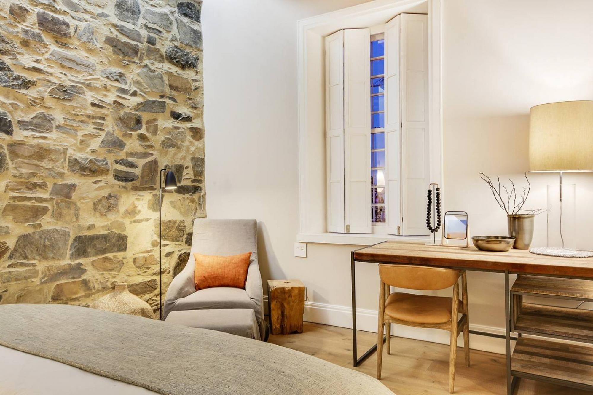 A room with a stone wall, a bed, a window with shutters, a desk with a chair, a lamp, and a small table with decorative items.