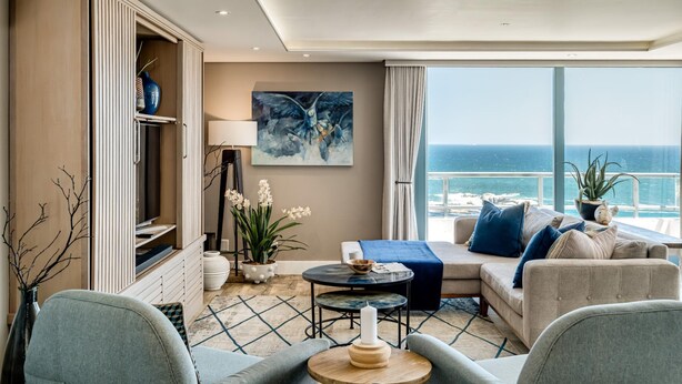 A modern living room with a sofa, a coffee table, and a TV. There is a large window with a view of the ocean. - Stylish Secure Camps Bay Holiday Apartment 9 Nautica (Cape Town)