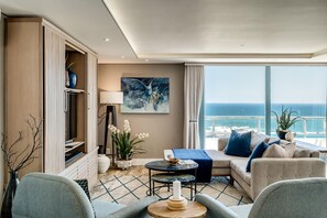 Uma sala de estar moderna com um sofá, uma mesa de centro e uma TV. Há uma janela grande com vista para o oceano.