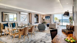 A spacious living area with a dining table, chairs, a sofa, and a large window offering a view of the sea. - Stylish Secure Camps Bay Holiday Apartment 9 Nautica (Cape Town)