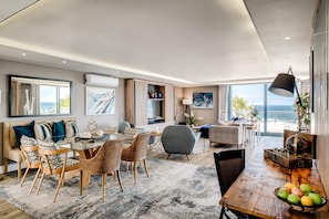 A spacious living area with a dining table, chairs, a sofa, and a large window offering a view of the sea. - Stylish Secure Camps Bay Holiday Apartment 9 Nautica (Cape Town)