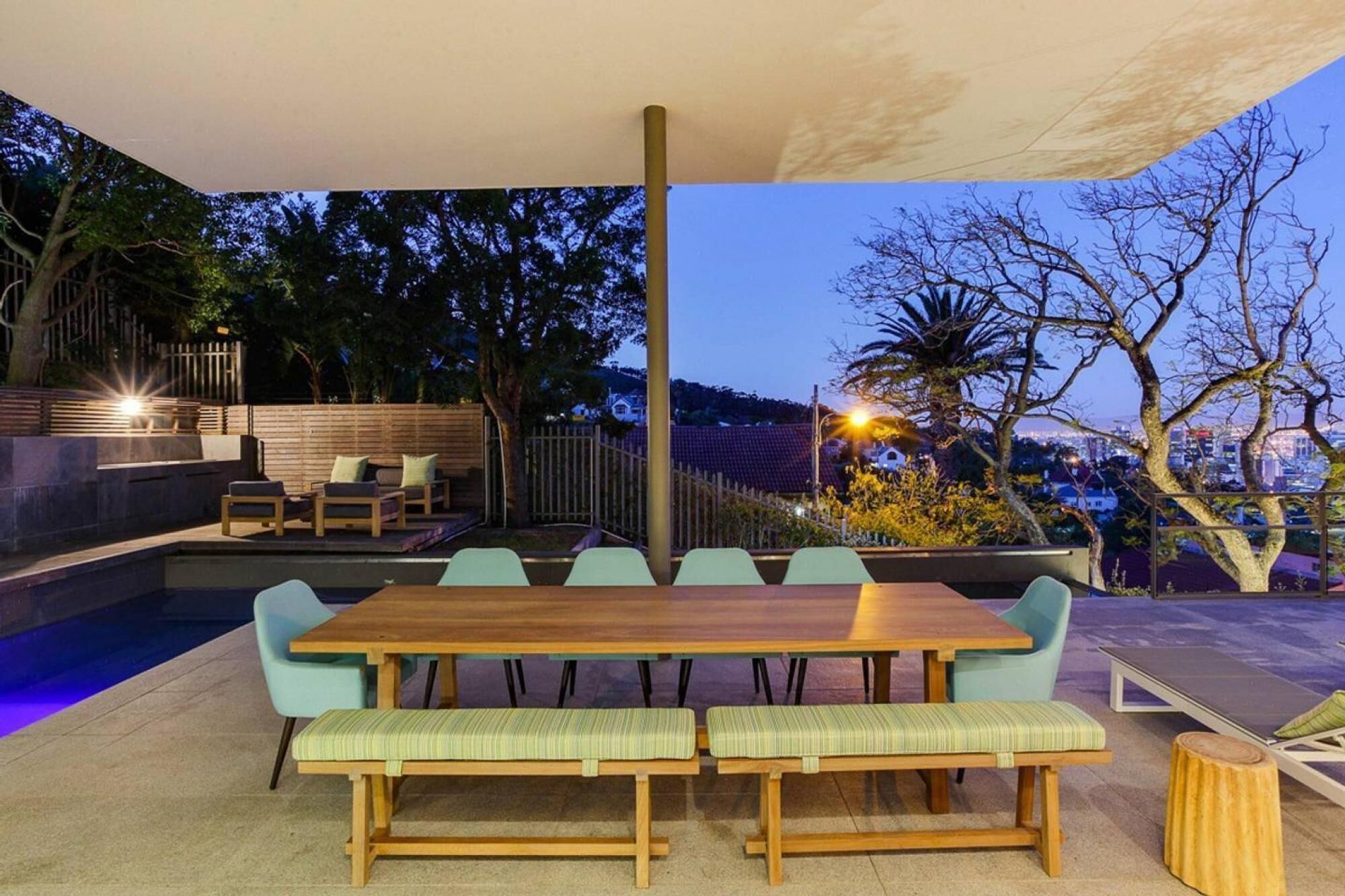 An outdoor dining area with a wooden table and benches, surrounded by a pool and trees.
