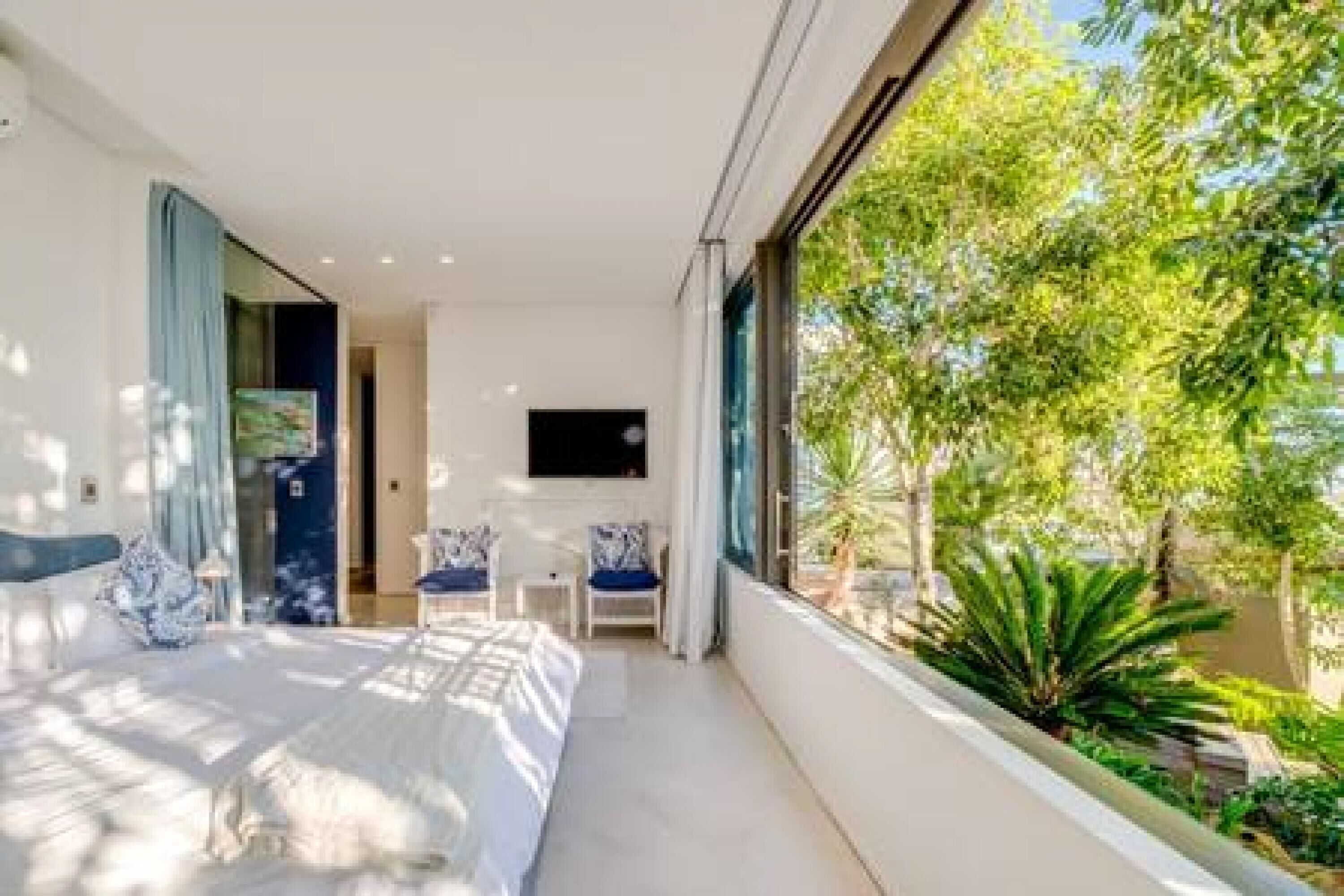 A modern bedroom with a large bed, a TV, and a view of greenery outside.