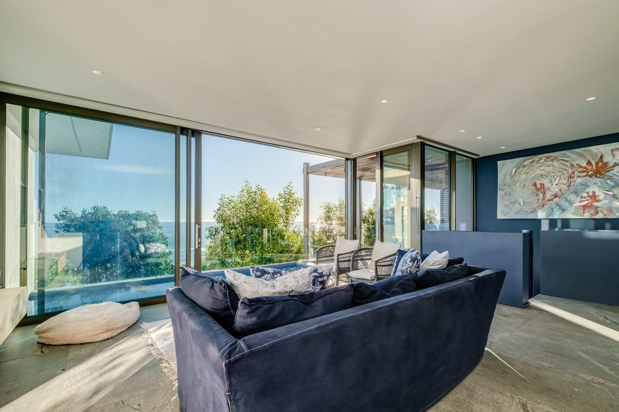 A modern living room with a large sofa, a dining area, and a view of greenery and the sea.