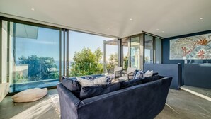 A modern living room with a large sofa, a dining area, and a view of greenery and the sea. - Unique Camps Bay Apartment w Private Pool Coral Sea (Cape Town)