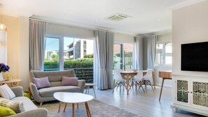 A modern living room with a sofa, a coffee table, a dining area with chairs, and a TV mounted on the wall. - Stylish 2 Bedroom Apartment in Camps Bay Asilvertide (Cape Town)