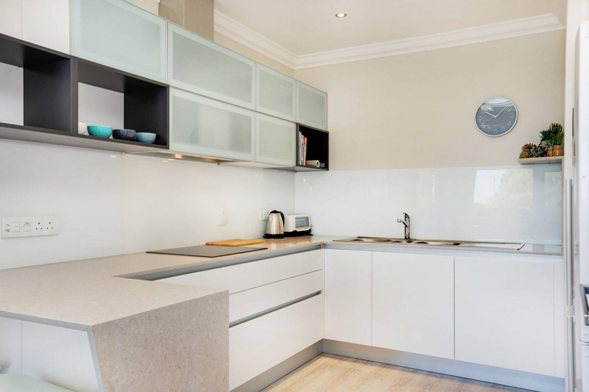 A modern kitchen with white cabinets, a built-in oven, a coffee maker, and a clock on the wall.