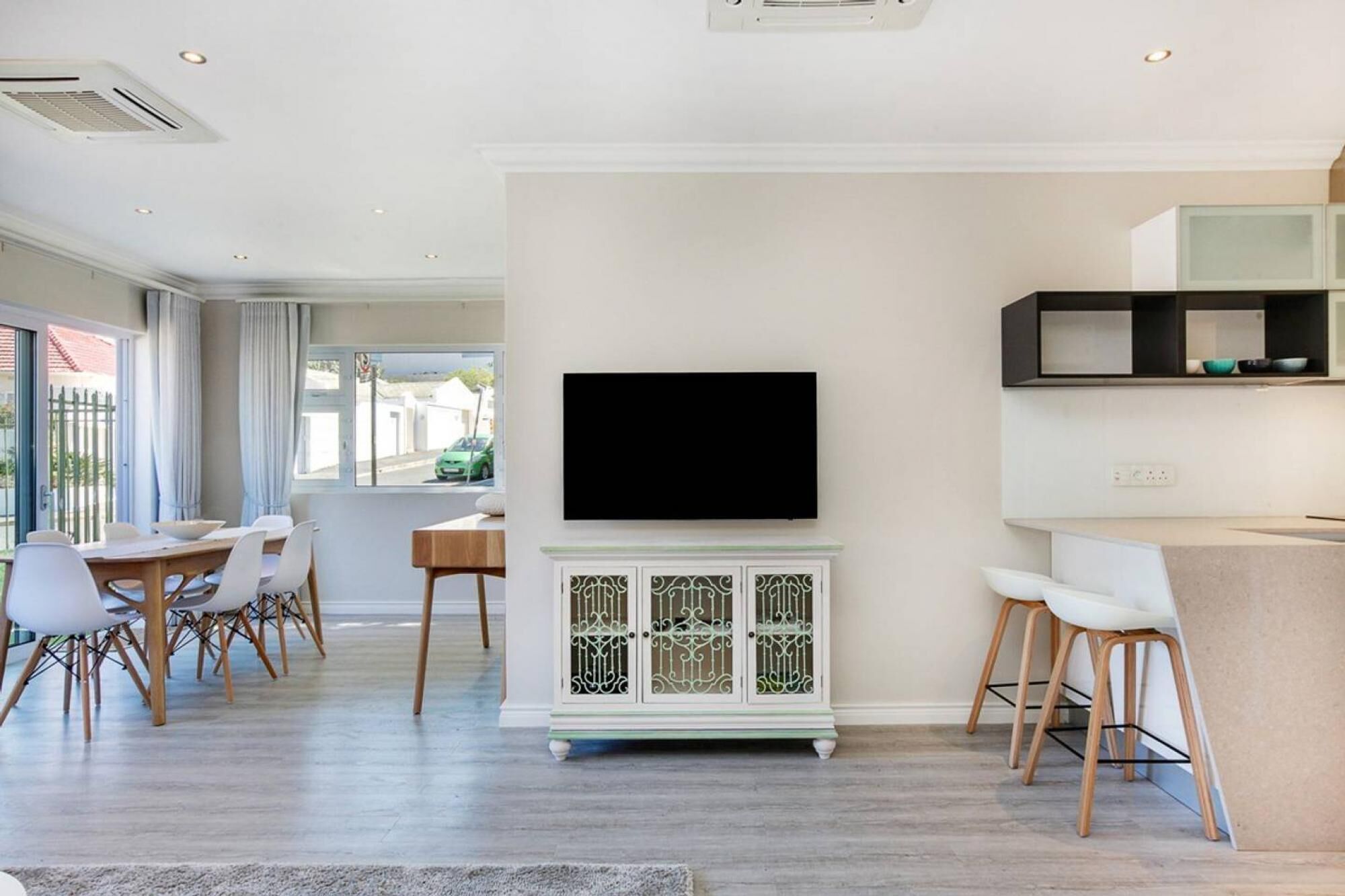 A modern living room with a flat-screen TV mounted on a white cabinet, a dining area with a wooden table and white chairs, and a kitchen area with a black countertop and white cabinets.