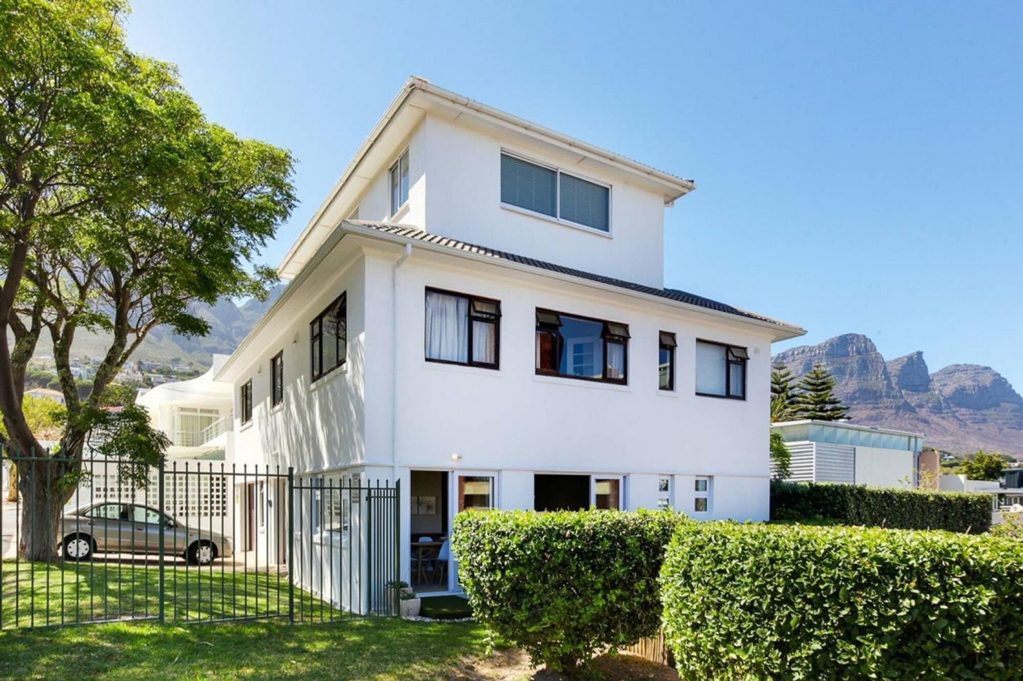 A two-story white house with a balcony, a car parked in the driveway, and a mountain in the background.